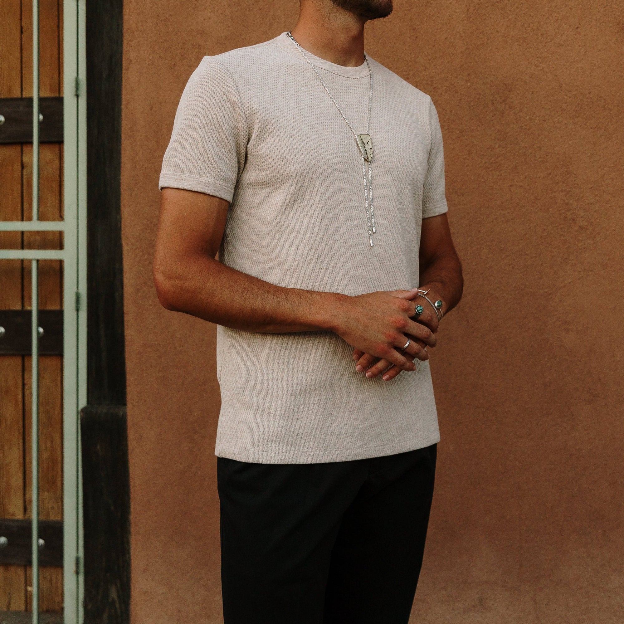 Man in beige knit tee wearing a silver pendant necklace and multiple rings.