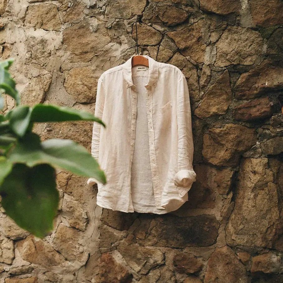 Vintage white linen Jack shirt with rolled sleeves hanging on a rustic stone wall beside green foliage.