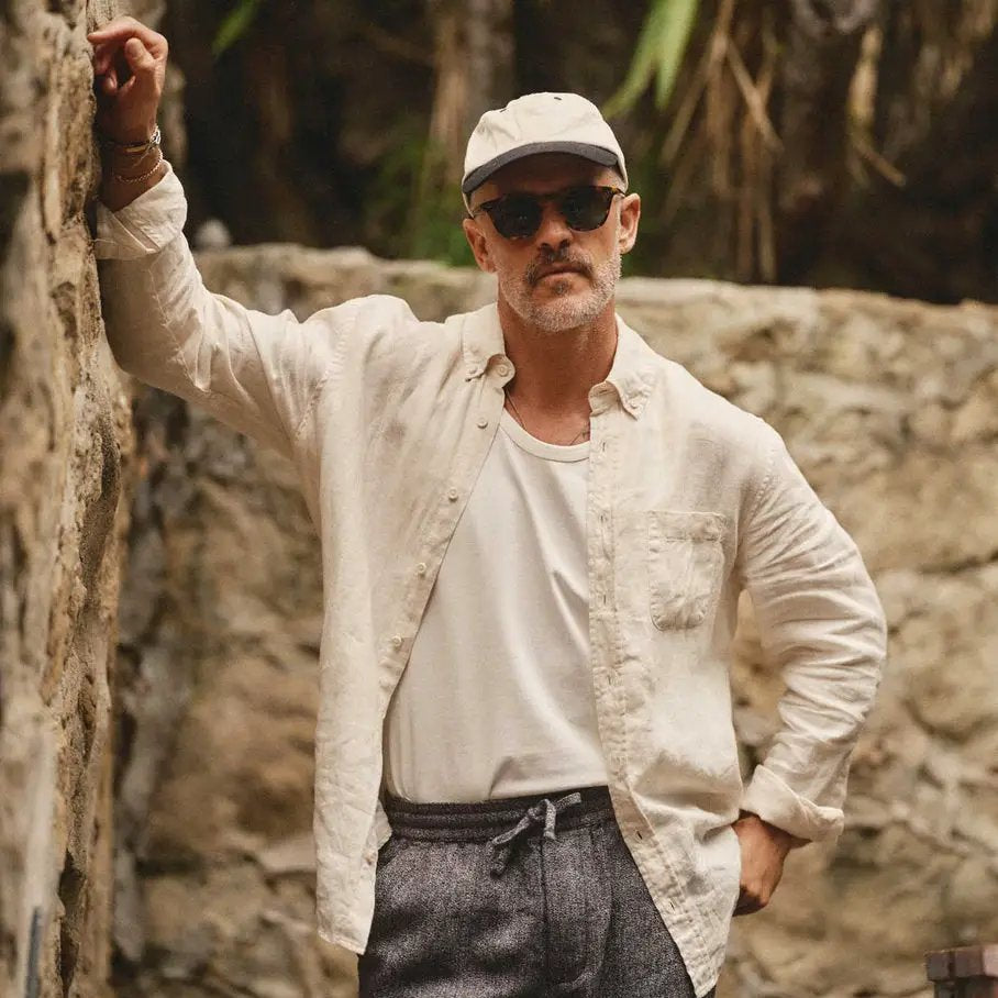Man wearing a vintage white linen shirt unbuttoned over a white tee, paired with grey drawstring pants and a cap against a stone wall.